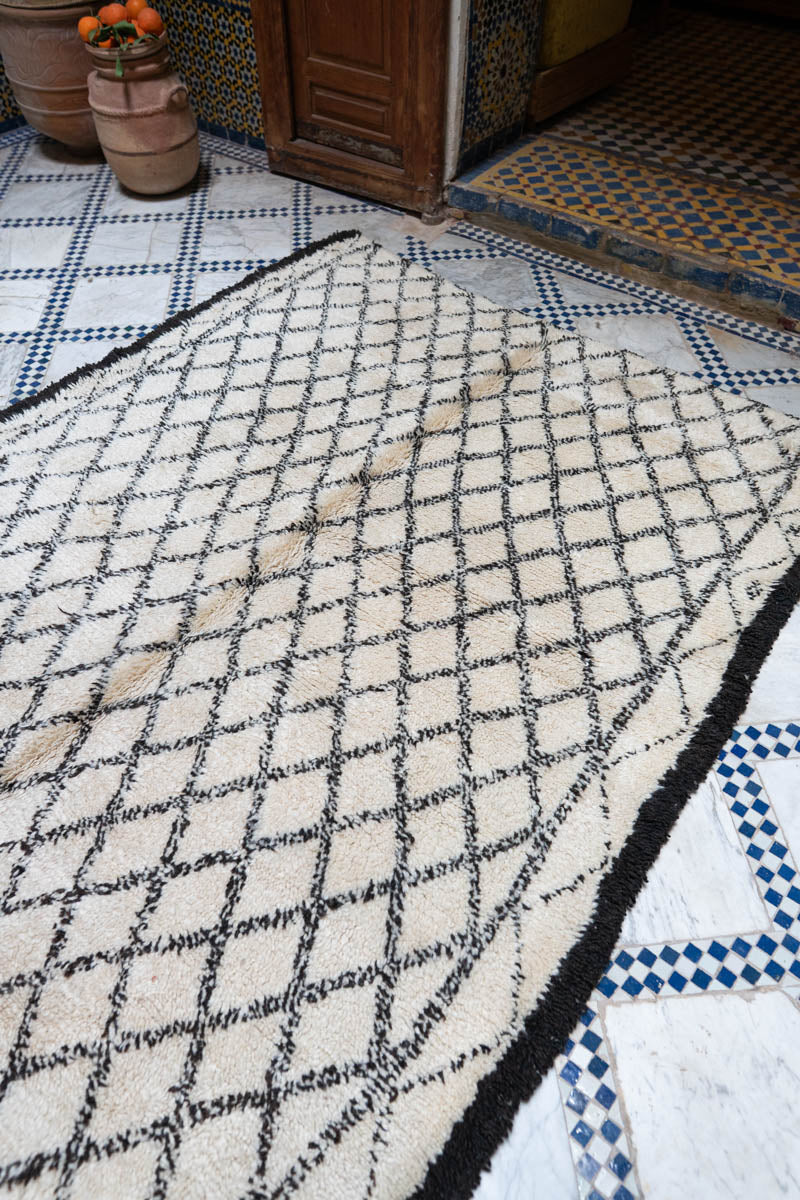 Pereinteinen marokkolainen riad, jossa  graafinen vintage-matto ja taustalla puiset ovet ja saviruukkuja