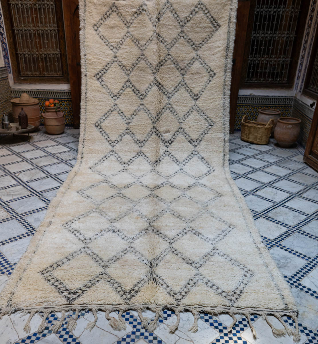 Beige and black patterned moroccan rug on a checkered floor with decorative pots in the background