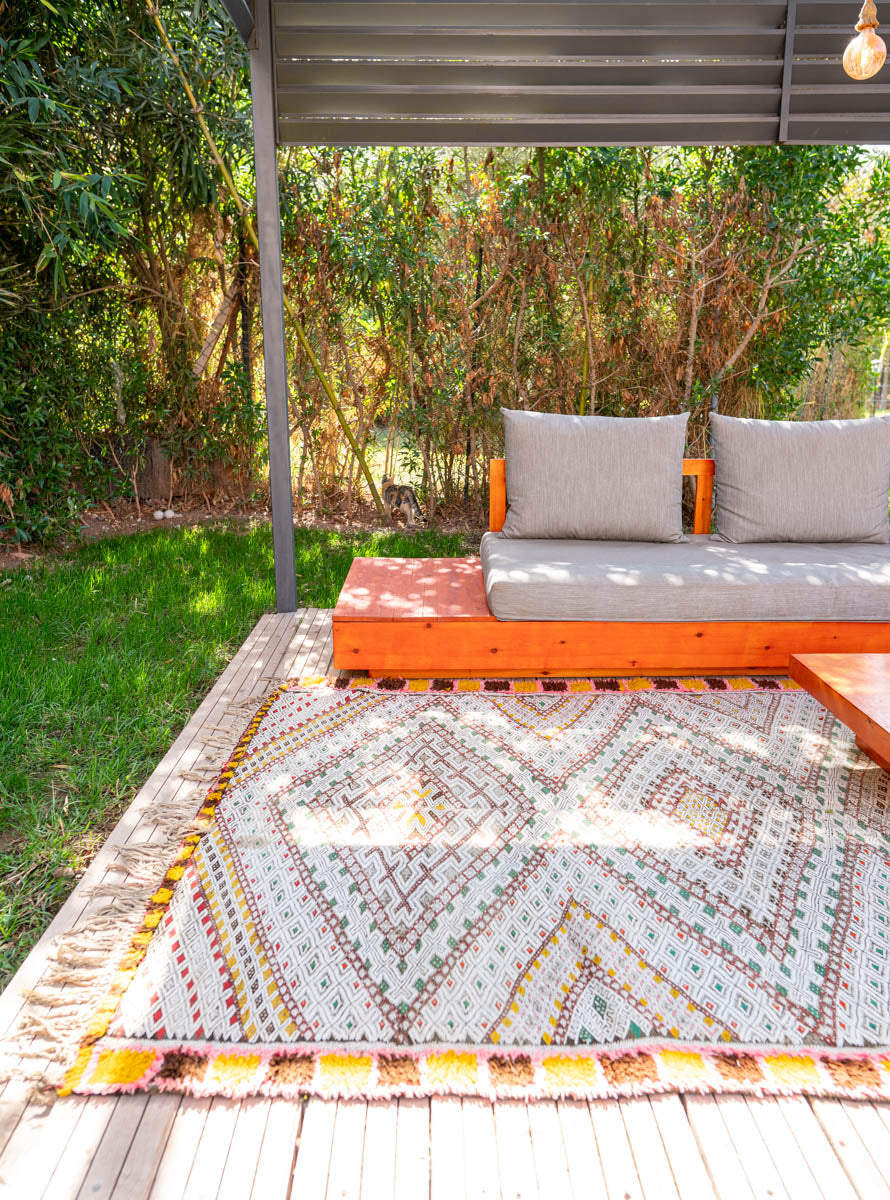 Outdoor seating area with a patterned moroccan kelim rug, orange bench, and gray cushions on a wooden deck.