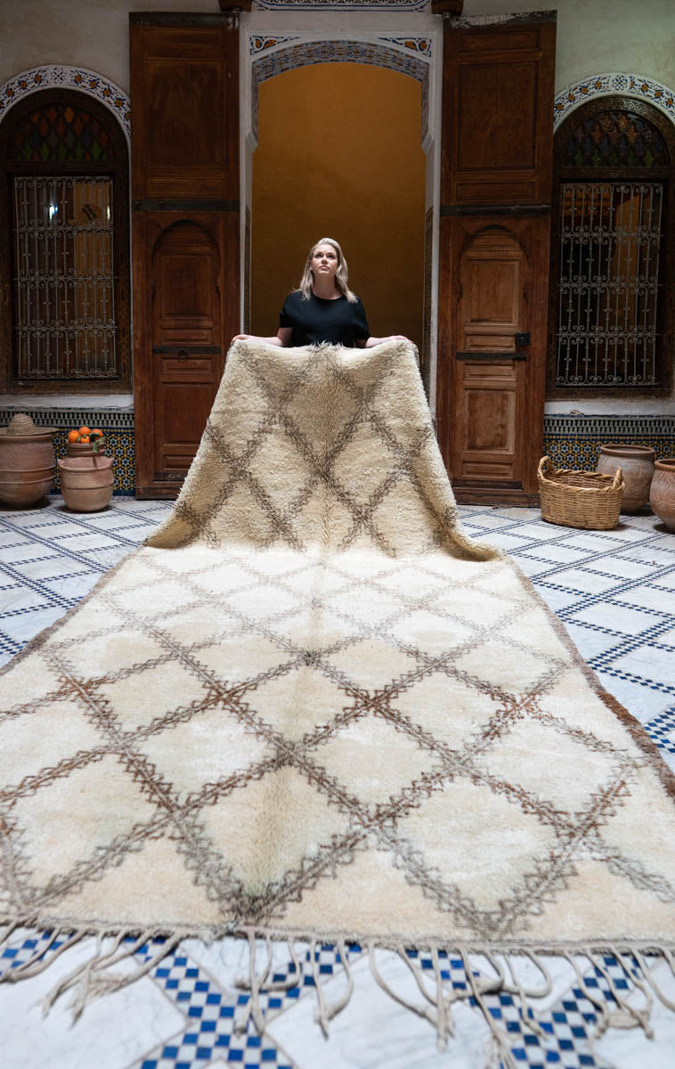 Woman standing behind a large woven Moroccan Beni Ouarain rug in a room with wooden doors and decorative elements.