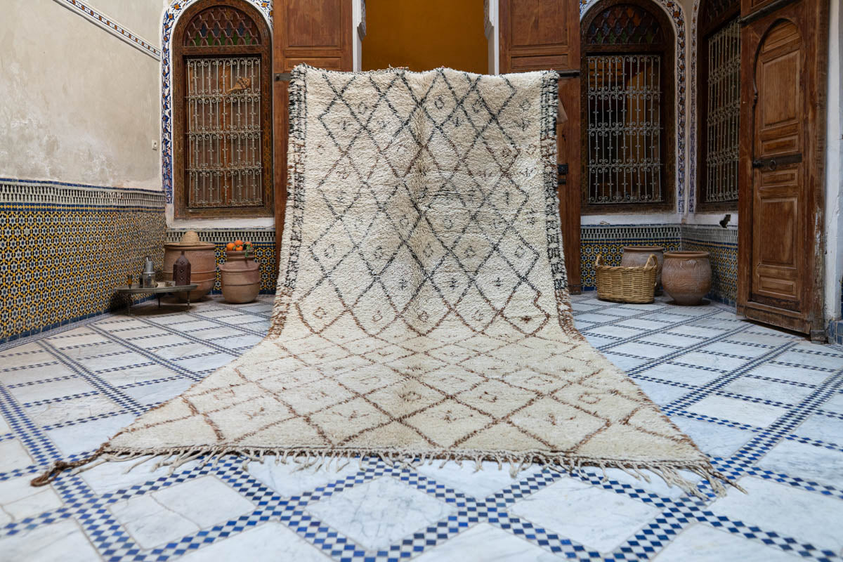 Koristeellinen marokkolainen vintage Beni Ouarain -matto geometrisella kuvioinnilla laattalattialla huoneessa, jossa on puiset ovet ja koriste-elementtejä.