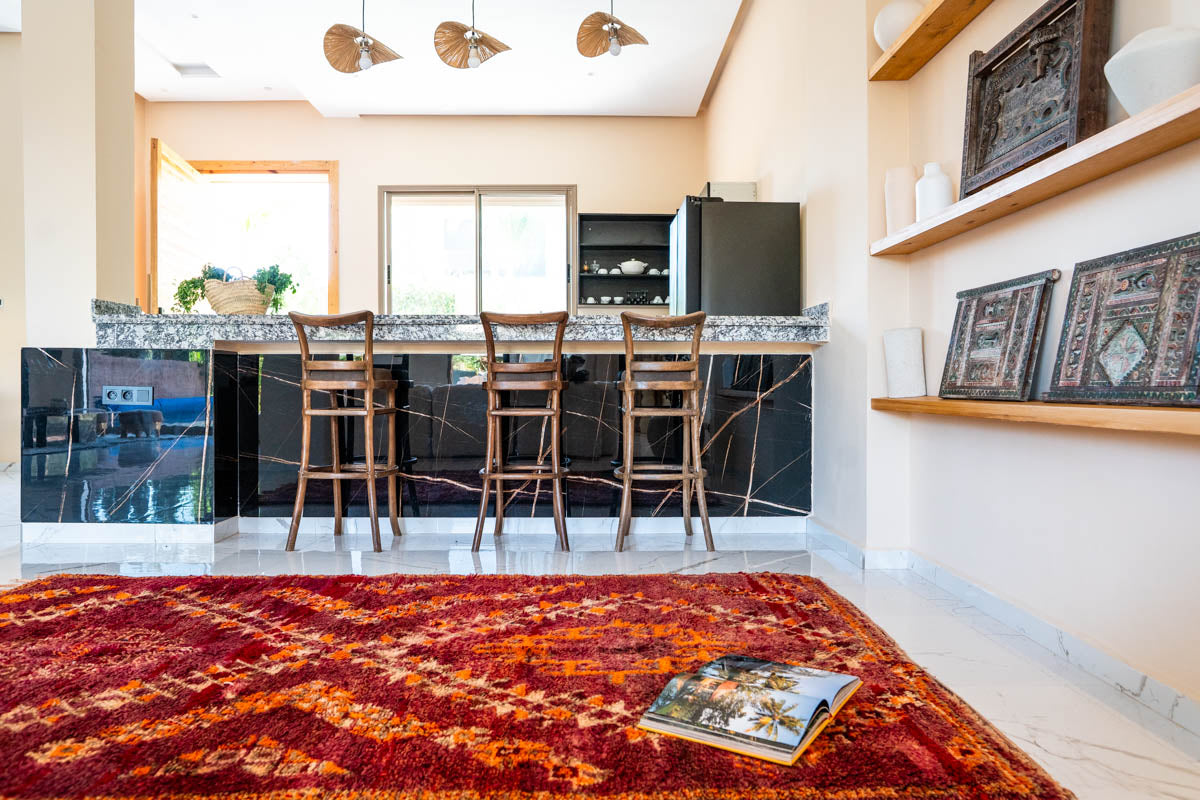 Modern kitchen with bar stools, a red patterned Moroccan rug, and decorative items on shelves.